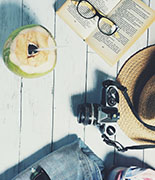 overhead shot of a coconut with a straw in it, an open book with a pair of reading glasses in the middle, a straw hat, a camera, a scarf, a pair of jeans with a pair of sunglasses on top. 