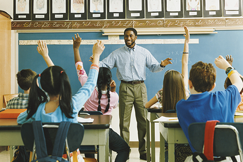 A teacher stands in front of a classroom filled with students who are raising their hands.